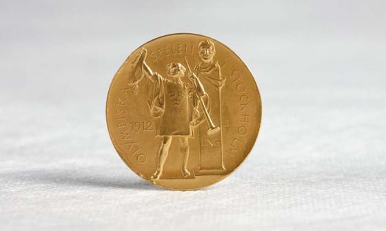 A gold medal standing upright on a white cotton surface. The medal shows a figure holding a trumped and a wine amphorae. Behind the figure is a bust on a stand. In raised lettering around the edge of the medal are the words 'Olympiska Spelen Stockholm' The year '1912' is also present.