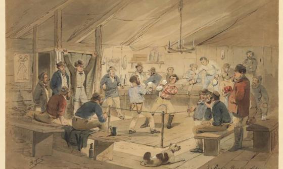 A group of approximately 20 men cheering and drinking whilst watching a boxing match. One boxer is in a blue shirt and one in a red shirt. A dog is in the foreground laying on the floor. 