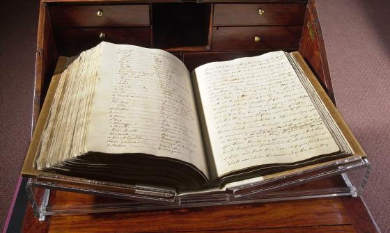 A large book open at a midway point. The paper is yellowed and stained. The writing is written in highly decorative cursive. The book is being held open on a stand which sits atop a richly varnished cabinet with draws and a fold out desk surface.