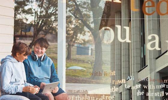 Two boys sit on a porch, smiling and looking at a tablet computer. The background is a blend of a rural scene and the National Library building.