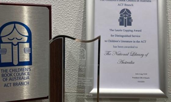 A silver and wooden plaque, glass trophy and framed certificate for the 2024 CBCA Laurie Copping Award given to the National Library