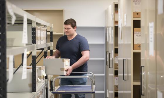 A person with short brown hair, a blue tshirt and jeans is organising archive boxes on metal shelves in a storage room. They are placing a box onto a shelf while standing next to a cart.
