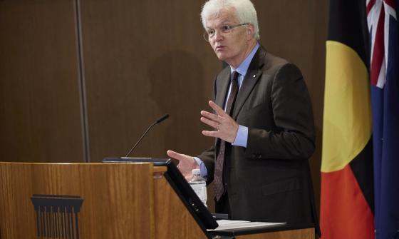 Professor Glyn Davis AC in a suit speaking at a podium and gesturing with his hands. Aboriginal and Australian flags are visible in the background.