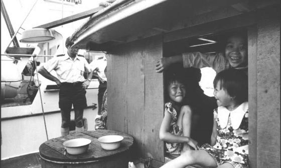 Three young girls smile and sit together inside a wooden boat cabin. A customs officer stands outside near a vessel labeled "Customs." Two bowls rest on a barrel near the cabin.