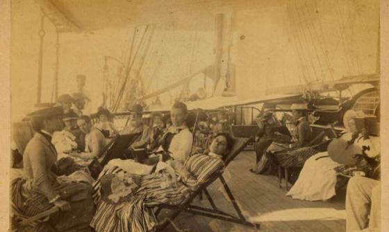 A sepia-toned photograph showing a group of passengers relaxing on the deck of a ship. Men and women are seated on deck chairs, dressed in late 19th or early 20th-century clothing, with hats and striped dresses visible. The background shows ship rigging and sails.