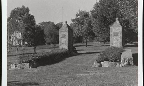 A black and white photograph of the main garrison gates of the former Cowra Prisoner of War camp. The gates now sit in a grassy park surrounded by trees and bushes.