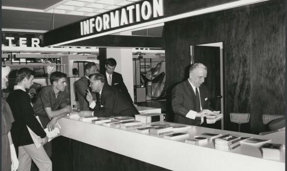 A black and white photograph of a group of men standing at a white information desk. Above the desk is a large sign that says INFORMATION. Men behind the counter and speaking and hanging out brochures.