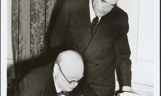 British Prime Minister Winston Churchill is seated at a table wearing glasses. He is signing a document. Standing behind him and leaning over his shoulder is Australian Prime Minister Robert Menzies. Both men are wear dark suits with white shirts. Menzies is wearing a dark tie, Churchill a bow tie.