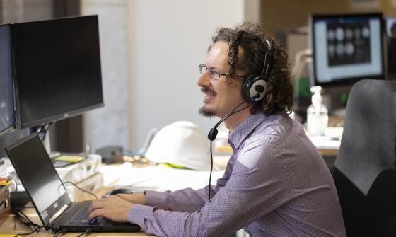 A person with curly hair, glasses and wearing a business shirt sits at a computer using a headset with microphone