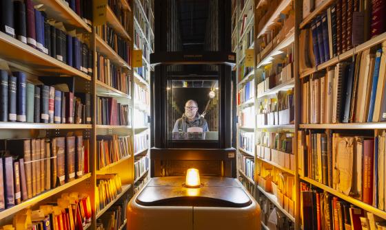 Person operating a lift between tall shelves filled with books, with a bright light at the front of the lift and warm overhead lighting.