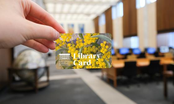 A hand holding a National Library of Australia library card featuring an image of yellow wattle flowers. The card is held in focus with a blurred background of the library's reading room, showing desks, chairs, and computer terminals.