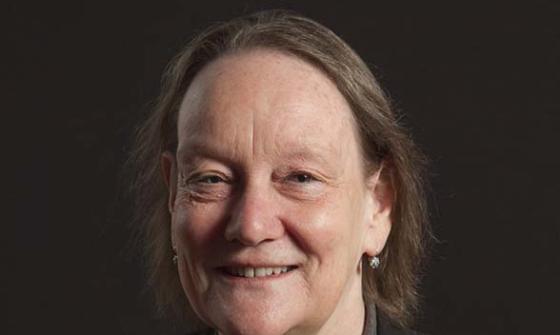 A portrait of a smiling woman wearing a black cardigan over a black top and a grey sweater. She is also wearing a colourful brooch shaped like a tree with colourful leaves.