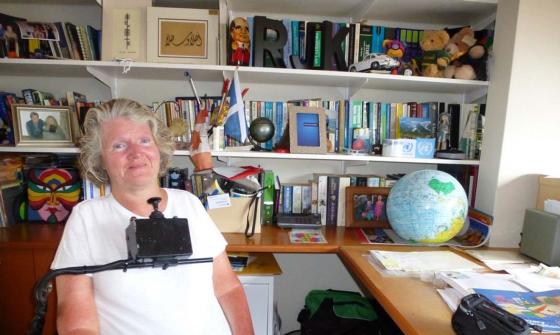 A photograph of a smiling woman seated in a wheelchair in front of a large bookcase and desk. She is wearing a white tshirt. The wheelchair has a control stick which sits just below her chin.