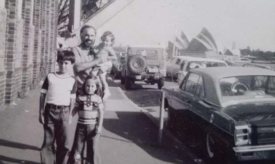 A black and white image of an asult and three children standing by the side of the road with an old fashioned car parked near them. The Sydney Opera House is in the background.