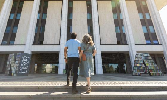 Man and woman in casual wear holding hands and laughing as they walk up the stairs towards the National Library entrance
