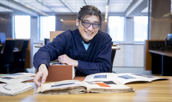 Dr Tets Kimura smiling and sitting at a table in the Special Collections Reading Room with material relating to Japanese fashion spread out in front of him
