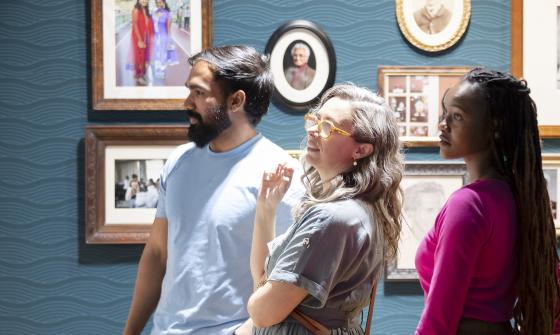Three people, one man and two women, looking at an item in the Exhibition Gallery at the Library. Behind them is a blue wall with framed family photos