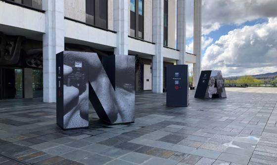 The front podium and colonnade of the National Library of Australia, taken on an angle. On the podium are three huge letters: an 'N', an 'L' and an 'A'. The letters have large photographs on them.