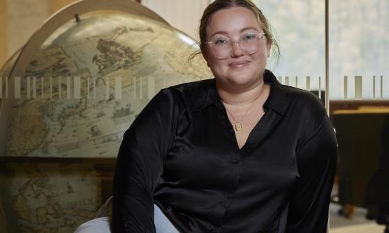 Woman, Jacinta Mackay, sitting in front of a window looking into the Main Reading Room with a large globe on the other side