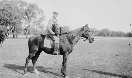 Black and white photo of a man with a large camera around his neck, sitting on a horse