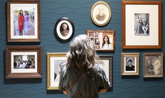 Woman with curly hair standing in front of a wall of framed family photos in the Hopes and Fears exhibition