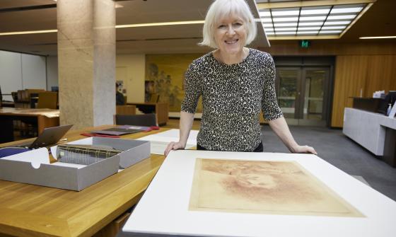 Woman, Dr Christina Thompson, standing in the Special Collections Reading Room, leaning on a table with a large portrait on it and smiling