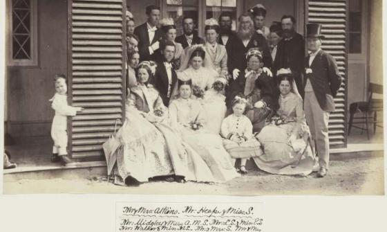 A black-and-white photograph showing a large wedding party posed on the steps of a wooden house. The group includes men, women, and children dressed in formal Victorian attire. The women are in elaborate dresses, some holding small bouquets. The men wear suits, some with top hats, and a small boy peeks around a door frame to the left.