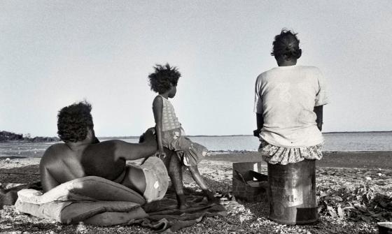 Black and white image of a man, woman and child sitting at a beach looking out towards the ocean