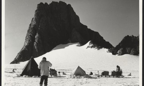 A man standing on a snowy plain, in front of tents, sledges with scientific equipment, scientists and sled dogs. A large rocky outcrop looms in the background. 