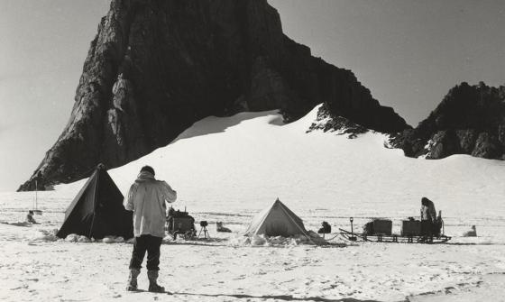 A man standing on a snowy plain, in front of tents, sledges with scientific equipment, scientists and sled dogs. A large rocky outcrop looms in the background. 