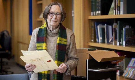 A woman with short hair and glasses smiles and looks at the camera while she holds a folded document in her hands. In the background are bookshelves with books and an open box on a trolley.