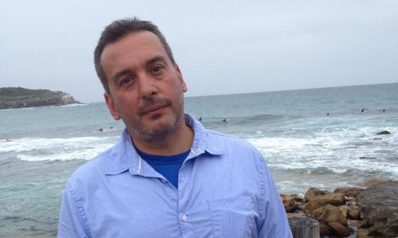 Christos Tsiolkas wearing a blue shirt standing in front of the ocean by a rock pool