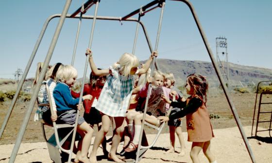 Photo of several young children in 1970s-era clothing playing on a swing set on a sunny cloudless day