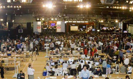 People standing, sitting at desks, and filming with news cameras in a brightly lit room
