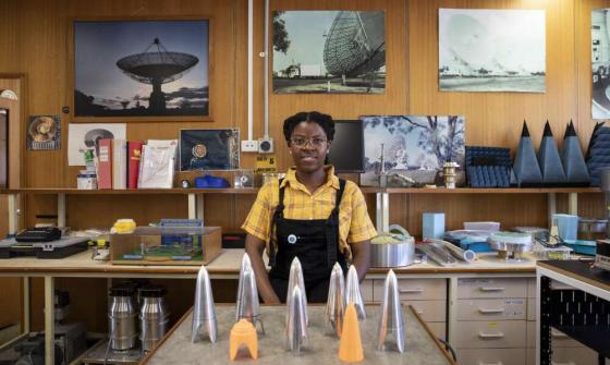 Natasha Maimbo, a young woman of colour, standing in a mechanical engineering lab with pictures of telescopes behind her. 
