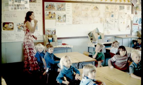 A colour photograph showing a teacher and children in a classroom in the 1970s