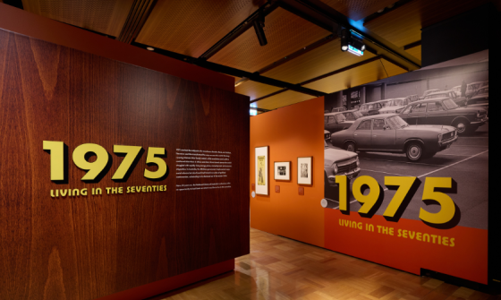 Entrance to the 1975: Living in the Seventies exhibition. A wall on the left is made to look like wood panelling with yellow text reading '1975: Living in the Seventies' and the wall on the right has a large black and white photo of 1970s-era cars 
