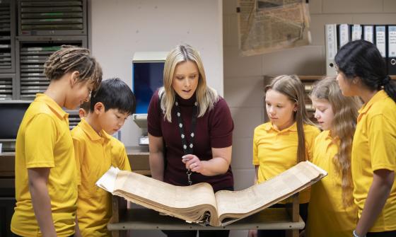 Five young school choldren standing around an old book with a Library staff member explaining the history of the book