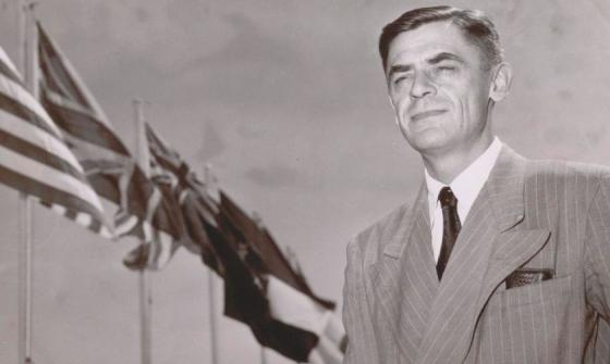 A black and white photograph of a man in a suit standing in front of flagpoles with the US and Australian flags