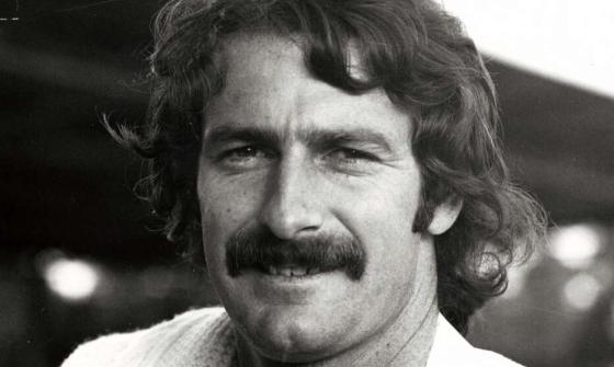 A black and white portrait of Dennis Lillee wearing cricket whites. A jumper is draped over his right shoulder.