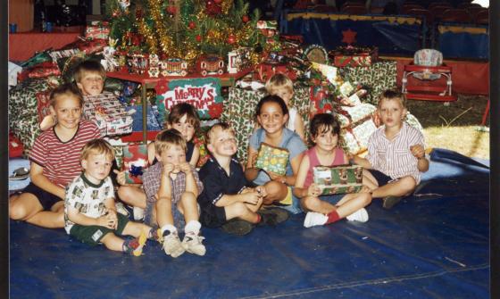 Group of 10 young children sitting under a Christmas tree with presents