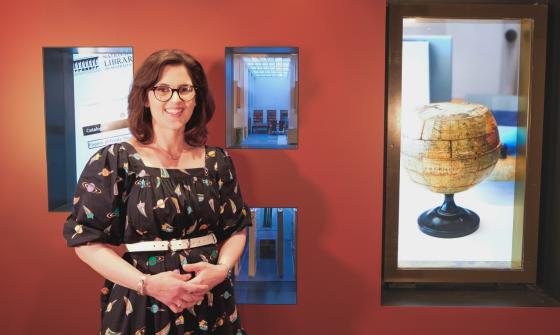 A dark-haired women wearing glasses stands, smiling, in front of a gallery display