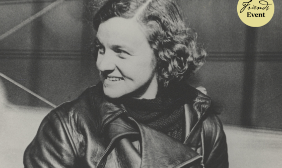 A woman stands in the centre of this image, looking to the left and smiling. She is in front of an aeroplane and wearing a jacket.