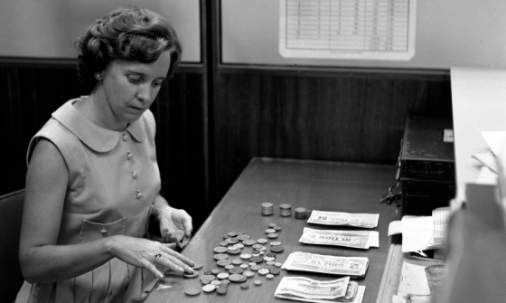 Black and white photo of a young woman counting coins and notes at a desk.