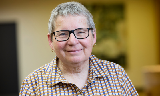 A short haired woman with black rimmed glasses in a checkered, button down shirt, smiling at the camera