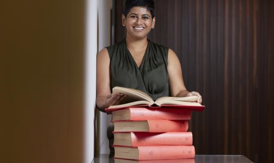 Natasha Naidu standing at a desk behind a stack of books holding one open and smiling at the camera