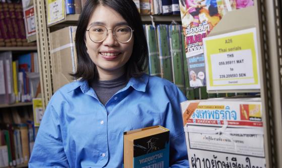 Woman with dark hair, Suprawee (Earn) Asanasak, standing in the stacks of the Library smiling and holding a book