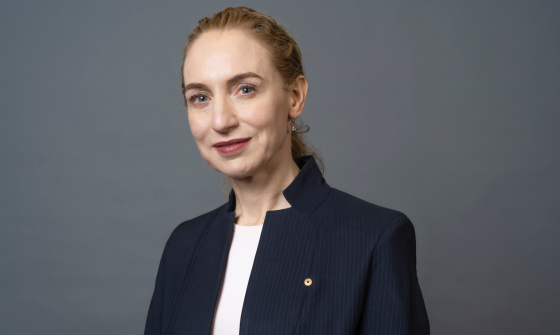 A woman with long hair tied back stands in the centre of the image wearing a white shirt and navy jacket