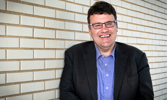 A man wearing a blue button down shirt and blazer smiling at the camera while standing in front of a white brick wall