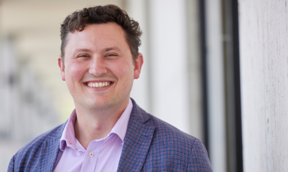 A man wearing a lilac button down shirt and a blue and red checkered blazer while smiling at the camera